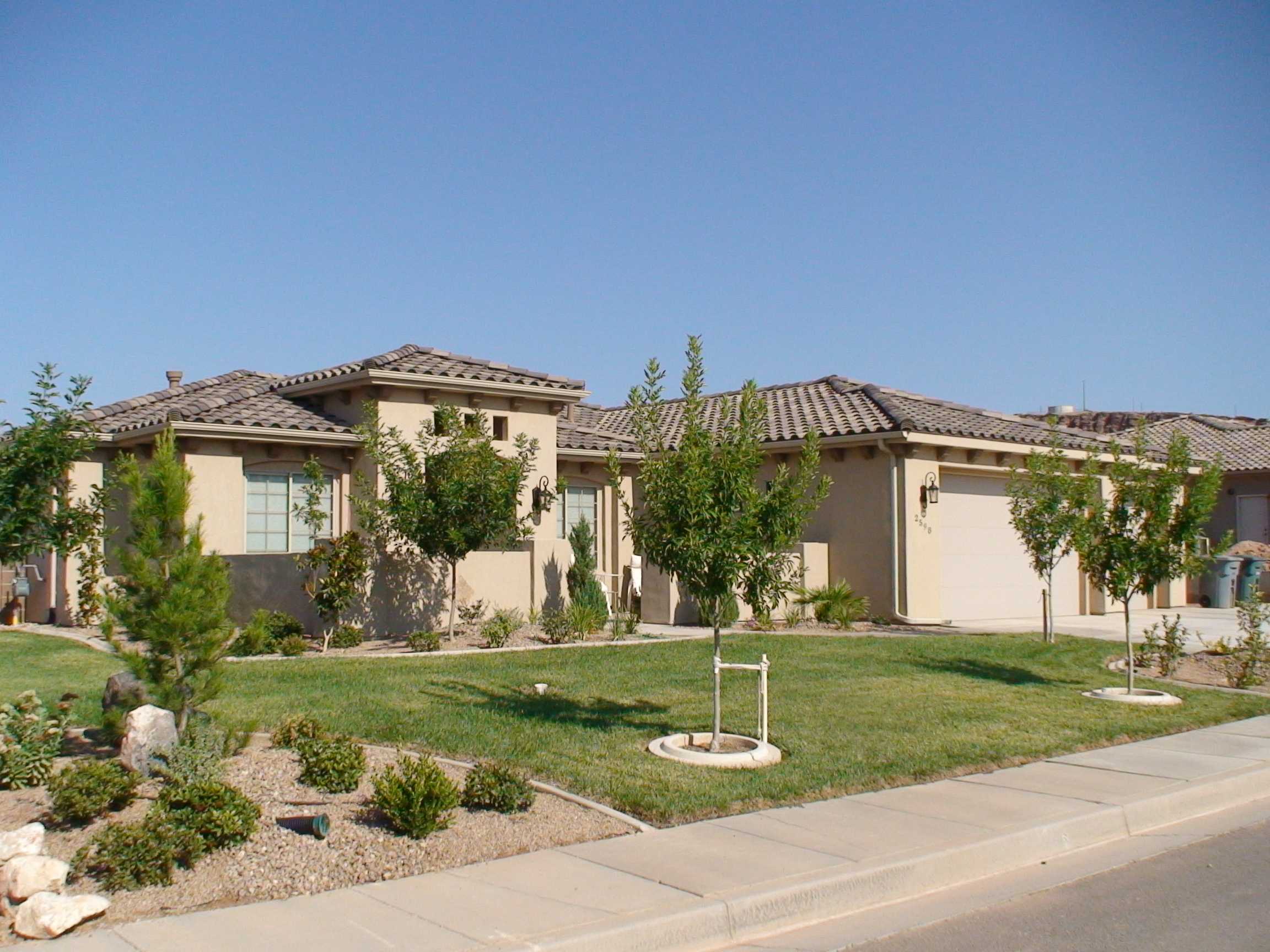 Single-story stucco house with a tiled roof, attached garage, and a landscaped front lawn with young trees and shrubs, set against a soft overcast sky in muted beige and green tones
